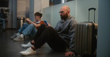 Kalabalık Uluslararası Havaalanı Terminali 'ndeki Çeşitli Yolcular: Bavullu Yorgun Adam Akıllı Telefon, Havayolu Merkezinin Kalkış Salonunda Oturuyor, Gecikmeli Uçuşu Bekliyor.