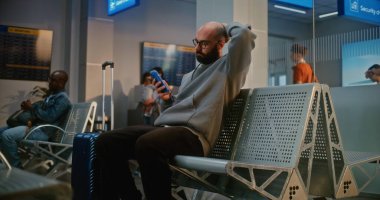 Kalabalık Uluslararası Havaalanı Terminali: Bavulu ve Belgeleri Olan Adam Güvenlik Kontrolünden Sonra Kalkış Salonunda Oturuyor, Telefonu Kullanıyor, Uçuşu Bekliyor. Pasaport Kontrolünde Duran İnsanların Kuyruğu.