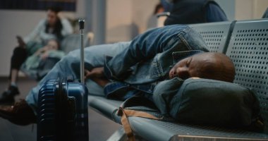 Kalabalık Uluslararası Havaalanı Terminali: Bavul ve Sırt Çantalı Yorgun Afro-Amerikan Turist Yataklı Havayolu Merkezinde Koltuklarda Uyuyor. Çeşitli Yolcular Gecikmeli Uçuş Bekliyor