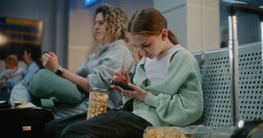 Kalabalık Uluslararası Havaalanı Terminali: Genç Kız Atıştırmalık Yiyen, Akıllı Telefon kullanarak Sosyal Medya Kaydıran, Uçak Uçuşunu Bekleyen. Havayolu Merkezinin Ayrılış Salonunda Farklı İnsanlar Oturuyor.