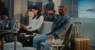 Kalabalık Uluslararası Havaalanı Terminali: Video Sohbeti 'nden Afro-Amerikan Adam Konuşuyor, Video İçeriği Akıllı Telefon Kullanıyor, Uçuşu Bekliyor. Havayolu İnbotu Biniş Salonundaki Çeşitli Yolcular.