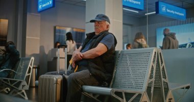 Kalabalık Havaalanı Terminali: Bavulu ve pasaportu olan yaşlı adam güvenlik kontrolünden sonra kalkış salonunda oturuyor, uçak biletini kontrol ediyor, uçuş için bekliyor. Belge Kontrolünde Duran İnsanların Kuyruğu.