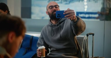 Kalabalık Uluslararası Havaalanı Terminali: Popüler Seyahat Blogcusu Akıllı Telefon Kullanarak Seyahat Video İçeriği, Uçuşu Bekleme. Havayolu Merkezinin Ayrılış Salonunda Farklı İnsanlar Oturuyor.