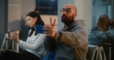 Kalabalık Uluslararası Havaalanı Terminali: Video Sohbeti 'nden Adam Konuşuyor, Trip' ten Video İçeriğini Çekiyor Akıllı Telefon Kullanıyor, Uçuşu Bekliyor. Farklı Yolcular Hava Yolları Merkezinin Kalkış Salonunda Oturuyorlar.