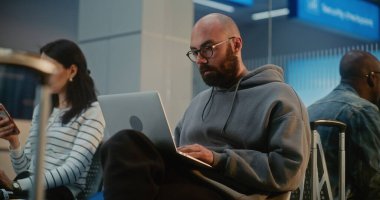 Kalabalık Uluslararası Havaalanı Terminali: Laptop 'ta Uzaktan Çalışan Adam, Çevrimiçi Görüntü Aranıyor, Uçak Bekliyor. Dijital Gadget 'ları kullanarak Havayolu İnbotu Biniş Salonundaki Çeşitli Yolcular.