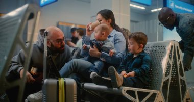 Kalabalık Havaalanı Terminali: İki Küçük Çocuklu Kadın Dijital Tablet 'te Çizgi Film İzliyor, Uçak Uçuşunu Bekliyor. Adam karısına kahve getiriyor. Havayolu Merkezinin Yatılı Salonunda Çeşitli İnsanlar