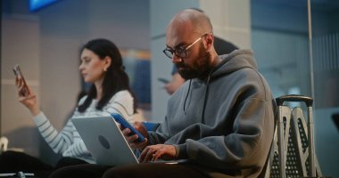 Kalabalık Uluslararası Havaalanı Terminali: İnternette Çalışan Girişimci, E-İş Yapıyor, Uçuşu Bekliyor. Dijital Aygıtları kullanarak, Hava Yolları Merkezinin Ayrılış Salonunda Oturan Çeşitli İnsanlar.