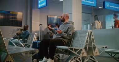 Kalabalık Uluslararası Havaalanı Terminali: Bavulu ve Belgeleri Olan Adam Güvenlik Kontrolünden Sonra Kalkış Salonunda Oturuyor, Telefonu Kullanıyor, Uçuşu Bekliyor. Pasaport Kontrolünde Duran İnsanların Kuyruğu.