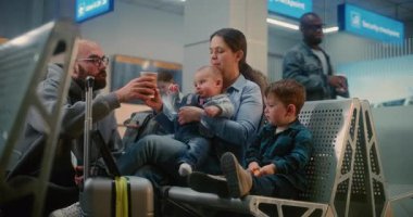 Kalabalık Havaalanı Terminali: İki Küçük Çocuklu Kadın Dijital Tablet 'te Çizgi Film İzliyor, Uçak Uçuşunu Bekliyor. Adam karısına kahve getiriyor. Havayolu Merkezinin Yatılı Salonunda Çeşitli İnsanlar