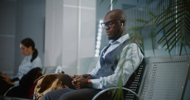 Uluslararası Havalimanı Terminali: Afro-Amerikan İşadamı Dijital Tablet ve Smartphone kullanarak, Uzaktan İşlem Yapıyor. Girişimci Ayrılış Salonunda Uçuş Bekliyor. Yavaş Hareket, Orta Çekim.