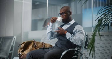 Havaalanı Terminali: Akıllı Telefon Kullanımı ve İçme Kahvesi ile Başarılı Girişimci. Afrika kökenli Amerikalı iş adamı, kalkış salonunda uçak bekliyor, iş gezisine çıkıyor. Yavaş Hareket, Orta Çekim.