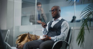 Havaalanı Terminali: Akıllı Telefon Kullanımı ve İçme Kahvesi ile Başarılı Girişimci. Afrika kökenli Amerikalı iş adamı, kalkış salonunda uçak bekliyor, iş gezisine çıkıyor. Yavaş Hareket, Orta Çekim.
