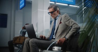 Uluslararası Havalimanı Terminali: Laptop Kullanımı ve İçme Kahve Kullanımı, E-İş Yapımı Başarılı Girişimci. Olgun İşadamı Uzaktan Çalışması Çevrimiçi Ayrılış Salonunda Uçuş Bekliyor. Yavaş Hareket.