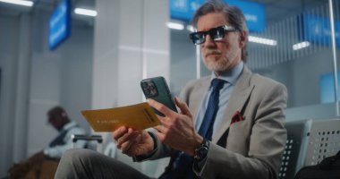 Havaalanı Terminali: Başarılı İşadamı Tarayıcı Uçuş Bilgilerini Kontrol ederek Akıllı Telefon kullanarak Uçuş Geçidi. Olgun Girişimci VIP Ayrılış Salonu 'nda oturuyor. E-Bilet Teknolojisi. Doli Vuruşu.