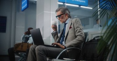 Uluslararası Havalimanı Terminali: Laptop Kullanımı ve İçme Kahve Kullanımı, E-İş Yapımı Başarılı Girişimci. Olgun İşadamı Uzaktan Çalışması Çevrimiçi Ayrılış Salonunda Uçuş Bekliyor. Yavaş Hareket.