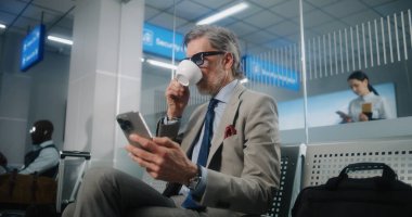 Uluslararası Havalimanı Terminali: Olgun İşadamı Ayrılış Salonu 'nda Oturmakta, Akıllı Telefon Kullanmakta ve Kahve İçmekte. Başarılı Girişimci Uçuşu Bekliyor, İş Gezisine Çıkıyor. Doli Vuruşu.
