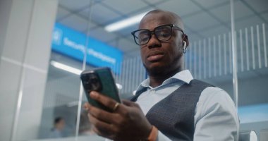 Havaalanı Terminali: Afrikalı Amerikalı Adam Akıllı Telefon kullanarak uçak biletini tarıyor, bilgileri kontrol ediyor. VIP Ayrılış Salonunda Uçağı Bekleyen İşadamı. E-Bilet Teknolojisi. Rack Odağını Kapat