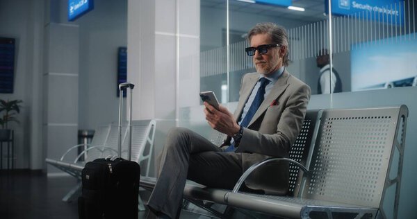 International Airport Terminal: Mature Businessman with Suitcase Using Smartphone, Sending Emails, Waiting for Flight. Entrepreneur Sitting in Departure Lounge of Airline Hub, Going on Business Trip.