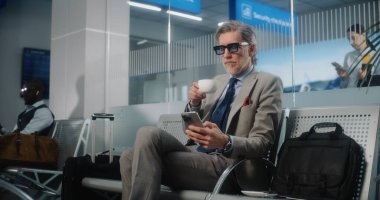 Havaalanı Terminali: Olgun İşadamı Akıllı Telefon Kullanıyor ve Kahve İçiyor, Gidiş Salonunda Bavulla Oturuyor. Girişimci uçuşu bekliyor, iş gezisine çıkıyor. Dolly shot, orta boy..