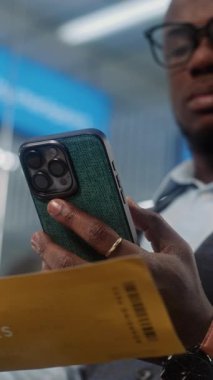 Havaalanı Terminali: Afrikalı Amerikalı adam akıllı telefon kullanarak uçak biletini tarıyor, bilgileri kontrol ediyor. VIP Ayrılış Salonunda Uçağı Bekleyen İşadamı. E-Bilet Teknolojisi. Dikey Görüntüyü Kapat
