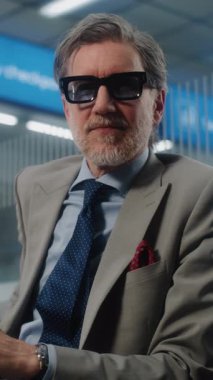 Havalimanı Terminali: Başarılı Gözlüklü Adam Kameraya Bakıyor, Akıllı Telefonu Tutuyor. Olgun İşadamı, Bavul Bekleme Salonu 'nda Uçuşu Bekliyor, İş Gezisine Çıkıyor. Dikey Çekim