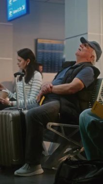 Kalabalık Uluslararası Havaalanı Terminali: Çeşitli İnsanlar ve Yolcular Gecikmeli Uçuşu Bekliyor, Seyahate Çıkıyor. Bavulu olan yaşlı adam, Ayrılış Salonunda Koltukta oturuyor. Dikey Çekim.