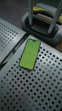 Havaalanı Terminalindeki Havayolu Hub Lounge: Yeşil Kroma Anahtarlı Akıllı Telefon Mockup Kahve ve Bavul 'un yanındaki koltukta uzanıyor. Sosyal Medya İçeriği için Yer Tutucu ile Görüntü Şablonu. Dikey Çekim.