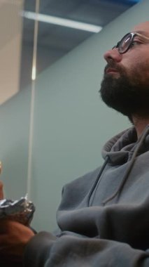 Kalabalık Uluslararası Havalimanı Terminali: Suitcase ile Yatılı Salonda Otururken Fast Food Burger Yiyen Adam. Çeşitli yolcular rötarlı uçuş bekliyor. El bilgisayarı. Dikey Çekim.