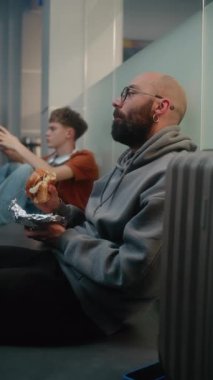 Kalabalık Uluslararası Havalimanı Terminali 'ndeki Çeşitli İnsanlar ve Yolcular: Burger Yiyen Adam, Yatılı Salonda Bavulla Oturan ve Gecikmiş Uçak Uçuşunu Bekleyen Adam. Dikey Çekim.