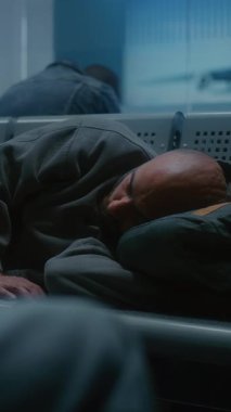 Kalabalık Uluslararası Havaalanı Terminali: Yorgun Adam Gece Gidiş Salonunda Sırt Sırtında Uyuyor. Çeşitli Yolcular ve Yolcular Gecikmiş Uçak Uçuşunu Bekliyor. Dikey Çekim.