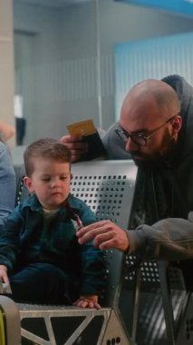 Kalabalık Havaalanı Terminalinde Uçuşu Bekleyen Çocuklu Aile. Kadın Bebekle Oynuyor, Erkek, Küçük Oğlu ile Dijital Tablet 'te Çizgi Film İzliyor. Airline Hub Lounge 'da çeşitli insanlar. Dikey Çekim