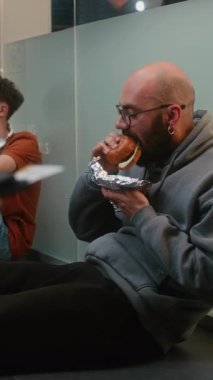 Kalabalık Uluslararası Havalimanı Terminali 'ndeki Çeşitli İnsanlar ve Yolcular: Burger Yiyen Adam, Yatılı Salonda Bavulla Oturan ve Gecikmiş Uçak Uçuşunu Bekleyen Adam. Dikey Çekim.