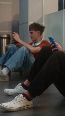 Kalabalık Uluslararası Havalimanı Terminali 'ndeki çeşitli yolcular: Çantalı Yorgun Adam Akıllı Telefon, Kalkış Salonunda Yerde Oturan, Gecikmeli Uçuşu Bekleyen. Dikey Çekim.