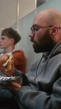 Kalabalık Uluslararası Havalimanı Terminali 'ndeki çeşitli yolcular: Adam Fast Food Burger yerken Ayrılış Salonunda Bavulla Oturuyor ve Gecikmeli Uçuşu Bekliyor. Dikey Çekim.