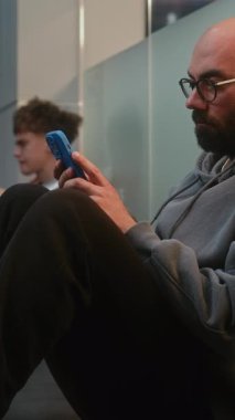 Kalabalık Uluslararası Havalimanı Terminali 'ndeki Çeşitli İnsanlar ve Yolcular: Yatılı Salonda Zeminde Oturan Yorgun Adam, Telefon Kullanıyor, Gecikmiş Uçak Uçuşunu Bekliyor. Dikey Çekim