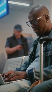 Kalabalık Uluslararası Havalimanı Terminali: Laptop kullanan Afrikalı Amerikalı Adam, Uzaktan Çalışıyor, Uçak Uçuşunu Bekliyor. Yatılı Salon 'da çeşitli turistler ve yolcular var. Dikey Çekim.