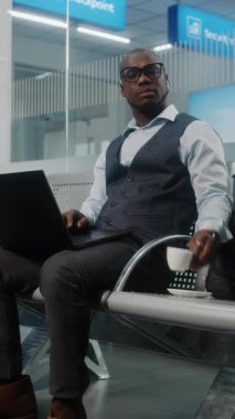 Havaalanı Terminali: Afro-Amerikan İşadamı Laptop Kullanıyor, Kahve İçiyor, Gülümsüyor ve Kameraya Bakıyor. Girişimci Uzaktan Çalışmalar Çevrimiçi Ayrılış Salonunda Uçuş Beklerken. Dikey Çekim
