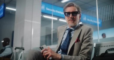 Havalimanı Terminali: Başarılı Gözlüklü Adam Kameraya Bakıyor, Akıllı Telefonu Tutuyor. Olgun İşadamı, Bavul Bekleme Salonu 'nda Uçuşu Bekliyor, İş Gezisine Çıkıyor. Doli Vuruşu.