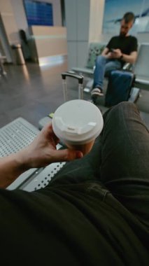Kalabalık Havalimanı Terminali: POV Yolcu Uçan Daire 'nin Yatılı Salonunda Oturuyor, Kağıt Kupadan Kahve İçiyor. Turistler uçuşu bekliyor, tatile çıkıyorlar. Birinci Şahıs Görünümü. Dikey Çekim