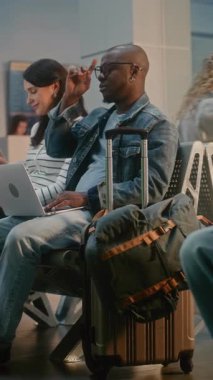 Kalabalık Uluslararası Havaalanı Terminali: Ayrılış Salonunda oturan çeşitli yolcular, Uçak Uçuşunu Bekliyor, Telefonları Kullanıyor. Uzaktan kumandalı bilgisayarda çalışan Afrikalı Amerikalı adam. Dikey Çekim.