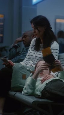 Anne ve Kızı Gece Gidiş Salonunda: Yorgun Ergen Kız Koltuklarda Yatıyor, Kadın Kaydırma Telefonu. Kalabalık Havaalanı Terminali 'nde çeşitli insanlar rötarlı uçuş bekliyor. Dikey Çekim