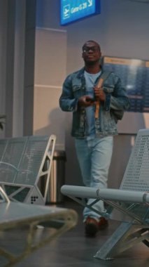 Havaalanı Terminali: Sırt çantalı Afrikalı Amerikalı adam güvenlik kontrolünden sonra bekleme salonunda oturuyor, telefonu kullanıyor, uçuşu bekliyor. Pasaport kontrolünde bekleyen yolcuların kuyruğu. Dikey Çekim.