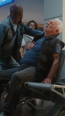 Kalabalık Havalimanı Terminali: Yaşlı Adam Kalp krizi geçiriyor, Infarction in Inarding Lounge of Airline Hub. Kıdemli Yolcuya Yardım Eden Çeşitli İnsanlar, Havaalanı İşçisi Tıbbi Yardım Çağırıyor. Dikey Çekim