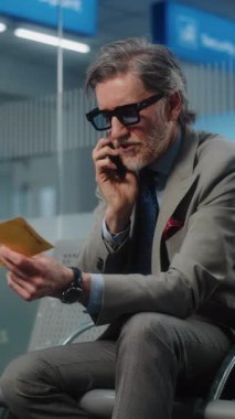 Havaalanı Terminali: Ciddi İşadamı Smartphone konuşuyor, Uçuş bilgilerini kontrol ediyor, uçak biletini tutuyor. Adam Ayrılış Salonu 'nda oturuyor, iş gezisine çıkıyor. Ağır çekim. Dikey Çekim.