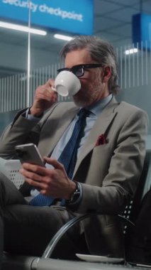 Havaalanı Terminali: Akıllı Telefon Kullanan ve Kahve İçen Olgun İşadamı, Kameraya Bakıyor. Girişimci, Bavul Ayrılış Salonunda Uçuş Bekliyor, İş Gezisine Çıkıyor. Dikey Çekim.