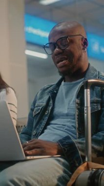 Kalabalık Uluslararası Havaalanı Terminali: Laptop 'ta çalışan Afro-Amerikan Adam, Çevrimiçi Video Görüşmesi Var. Çeşitli İnsanlar, Kalkış Salonunda oturan, Uçuşu Bekleyen Yolcular. Dikey Çekim.