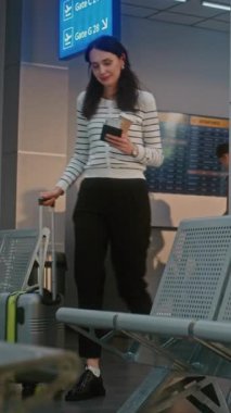 Kalabalık Havaalanı Terminali: Bavullu Kadın Güvenlik Kontrolünden Sonra Biniş Salonunda Oturuyor, Uçak Biletlerini ve Belgelerini Kontrol Ediyor, Uçuşu Bekliyor. Pasaport kontrolündeki yolcular. Dikey Çekim.