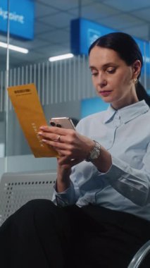 Havaalanı Terminali: Başarılı İşkadını Uçuş Bilgileri Kontrol, Akıllı Telefon kullanarak Uçuş Kartı Tarayıcısı. Kadın Girişimci VIP Ayrılış Salonu 'nda oturuyor. E-Bilet Teknolojisi. Dikey Çekim
