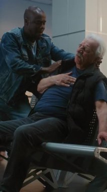 Kalabalık Havalimanı Terminali: Yaşlı Adam Kalp krizi geçiriyor, Infarction in Inarding Lounge of Airline Hub. Kıdemli Yolcuya Yardım Eden Çeşitli İnsanlar, Havaalanı İşçisi Tıbbi Yardım Çağırıyor. Dikey Çekim