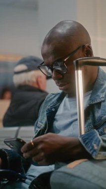 Havaalanı Terminali: Afro-Amerikan Bavulu Oturan ve Uçağı Bekleyen, Akıllı Telefon kullanan. Kalabalık Havayolu Merkezinin Ayrılış Salonu 'ndaki Çeşitli İnsanlar ve Yolcular. Dikey Çekim.